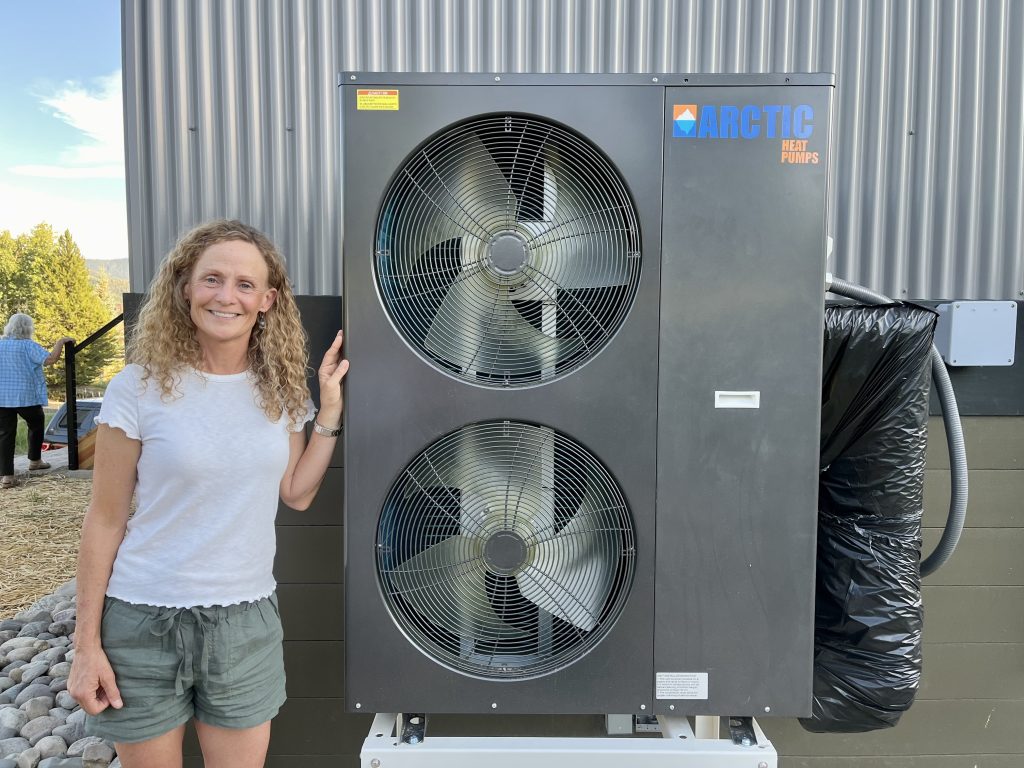 A woman stands next to a heat pump outside her home.