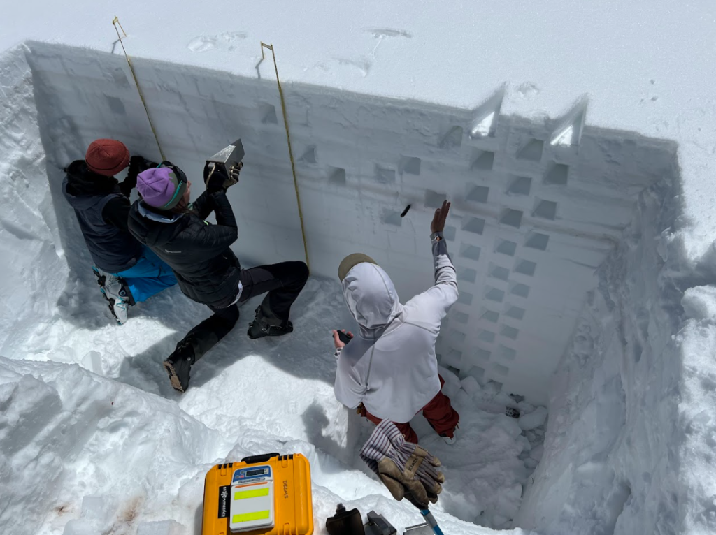 Three people collect snow density and temperature measurements in a snow pit.