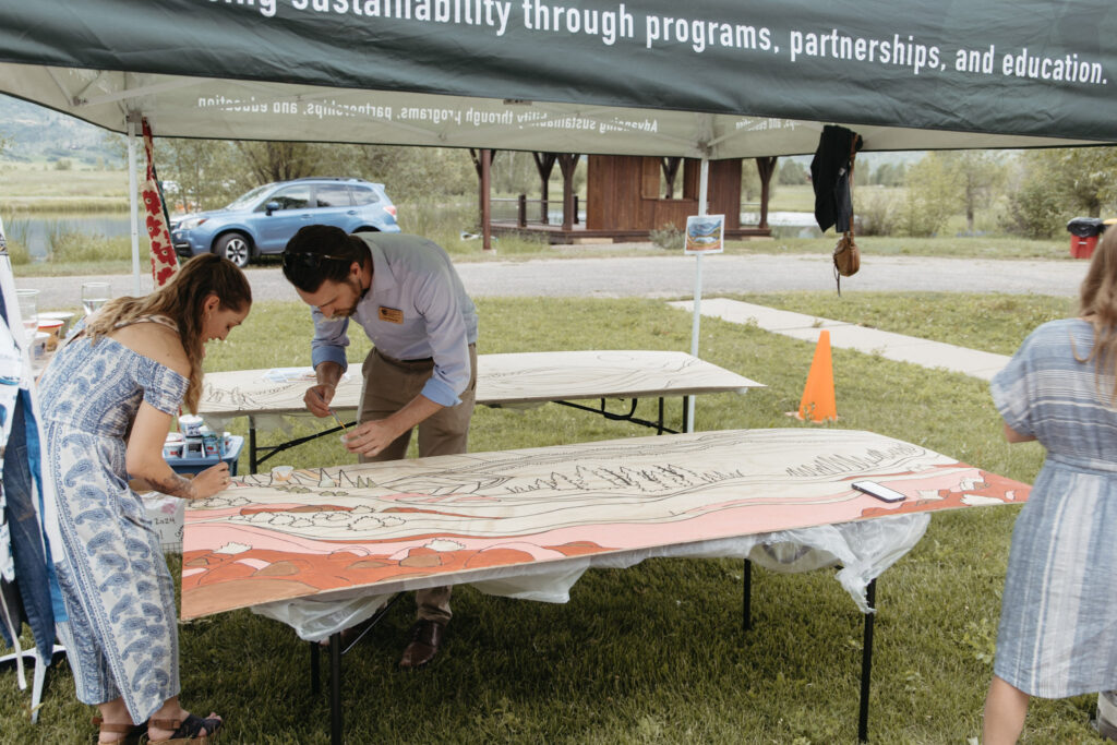 People stand outside over a wooden mural with paint brushes.