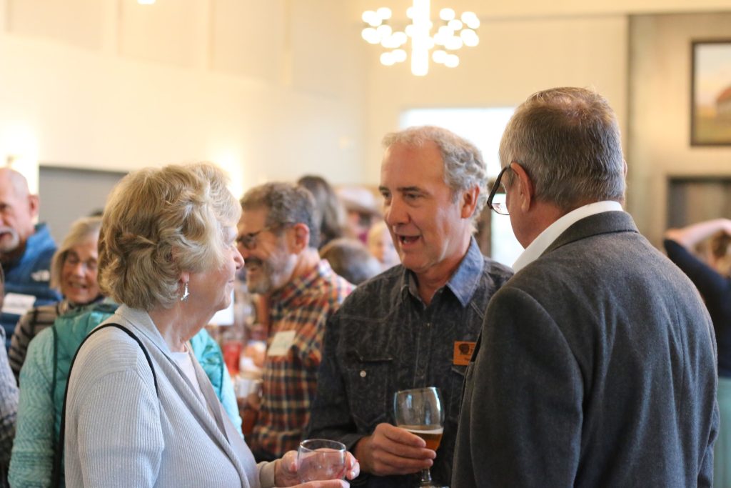 A man stands speaking to two people in a crowded room.