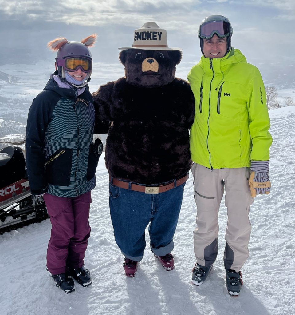 Two people post in ski gear on top of the mountain with Smokey the Bear.